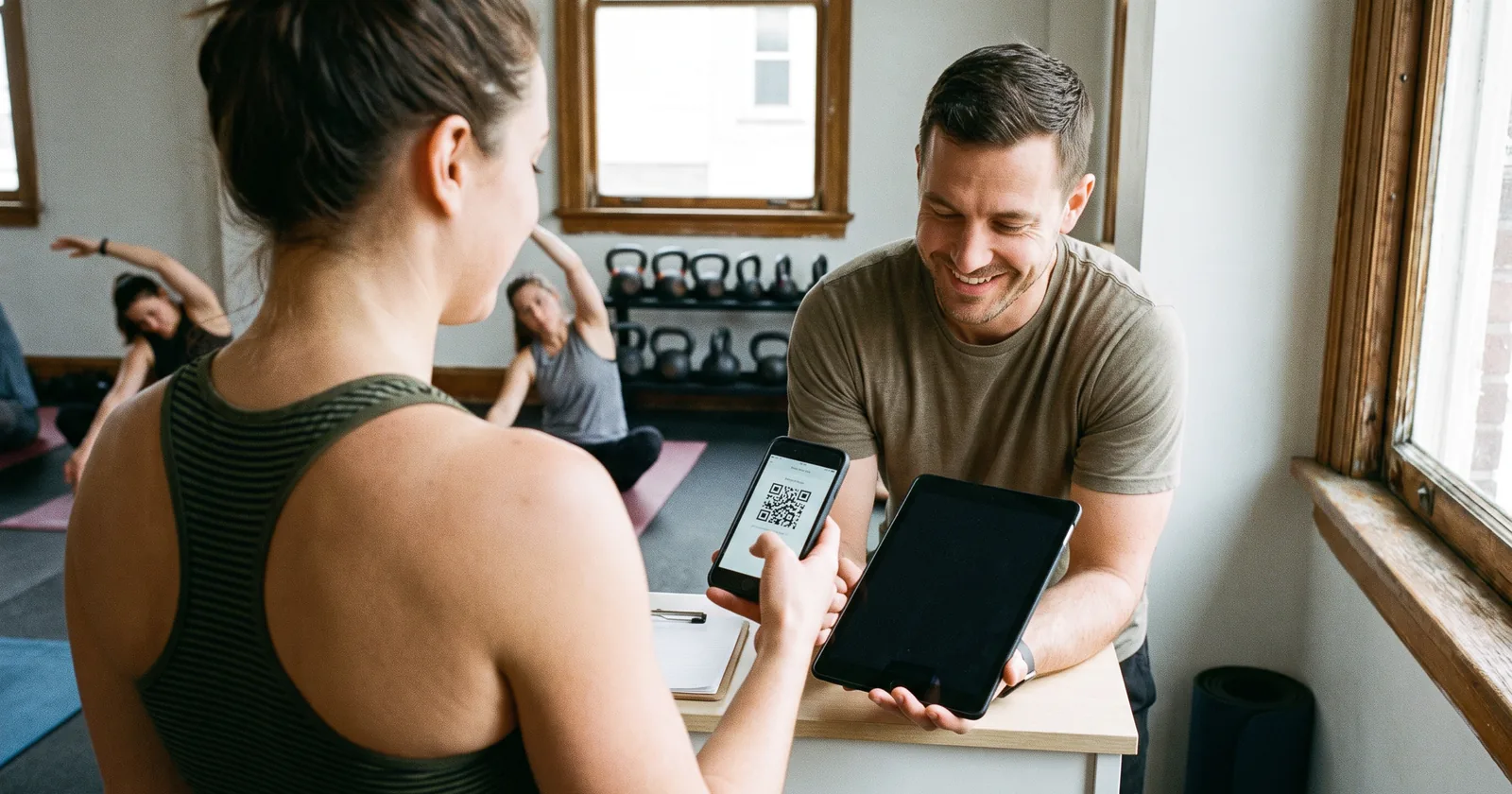 At a small fitness studio's front desk, a member in her club's colours shows the QR code for her fan deal while a class stretches behind her