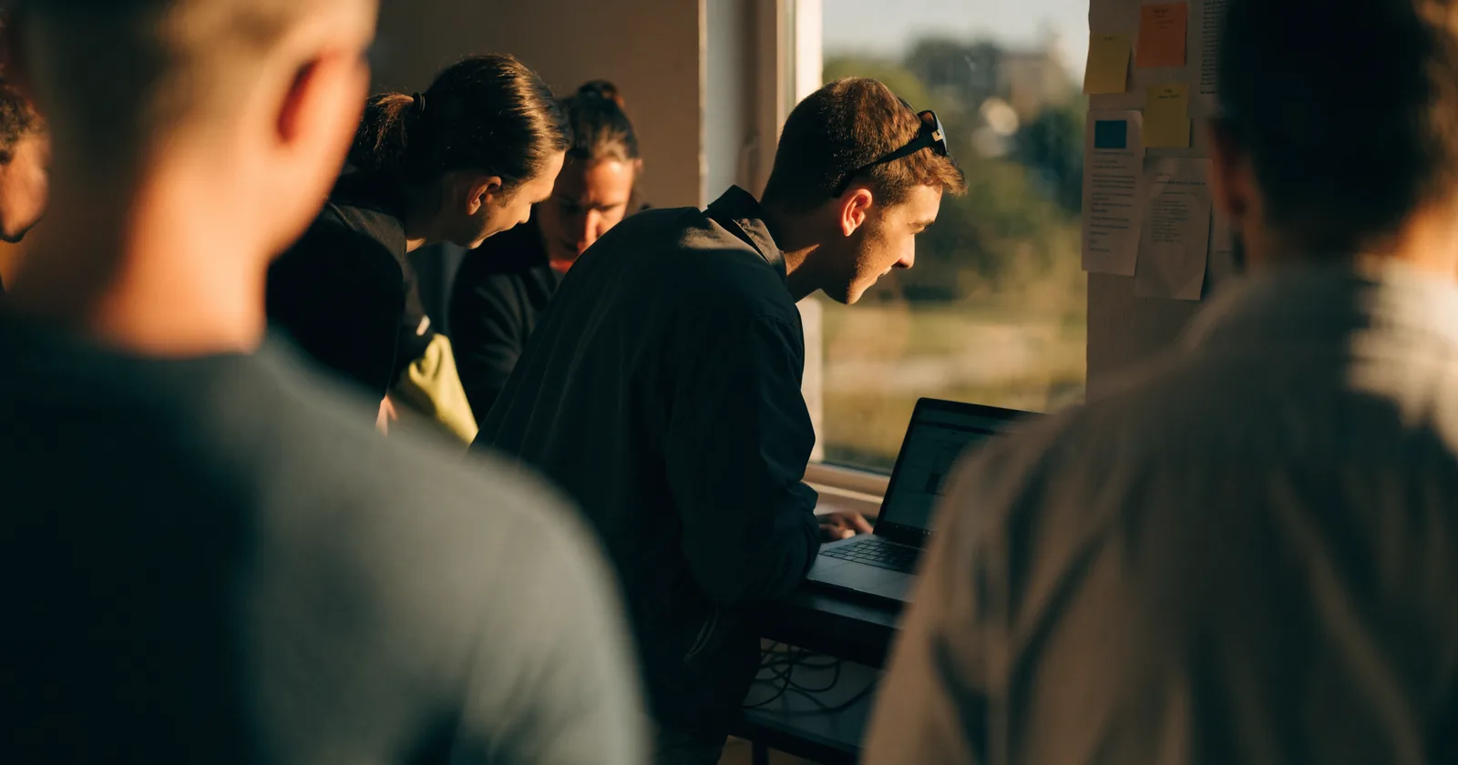 Small club staff around one laptop in a clubhouse office, late-afternoon light